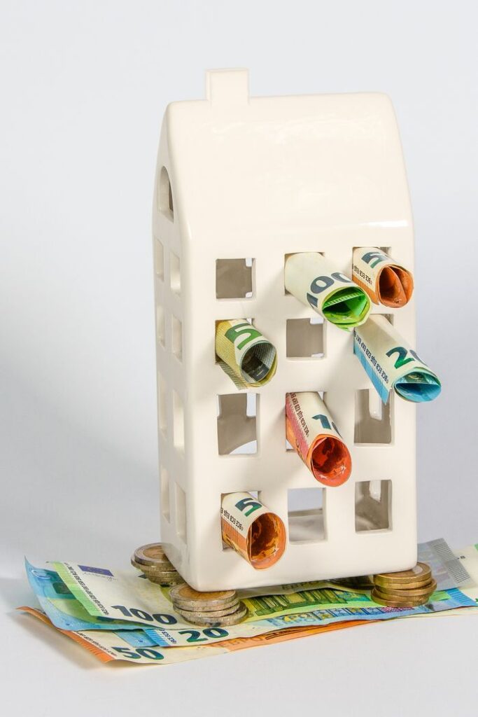 Ceramic house piggy bank stuffed with rolled euro notes and stacked coins, symbolizing real estate investment and savings.